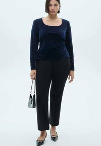 Marineblauwe longsleeve top met een vierkante halslijn, gecombineerd met zwarte op maat gemaakte broek. Model houdt een kleine zwarte handtas vast en draagt puntige ballerina's.