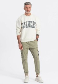 Mężczyzna z siwymi włosami i brodą, ubrany w białą bluzę z napisem "Los Angeles", oliwkowe spodnie cargo, białe sneakersy i czarny zegarek na ręku.