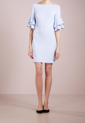 Femme portant une robe bleu clair mi-longue avec des manches à volants superposés et des chaussures noires à bout pointu, debout devant un fond uni.