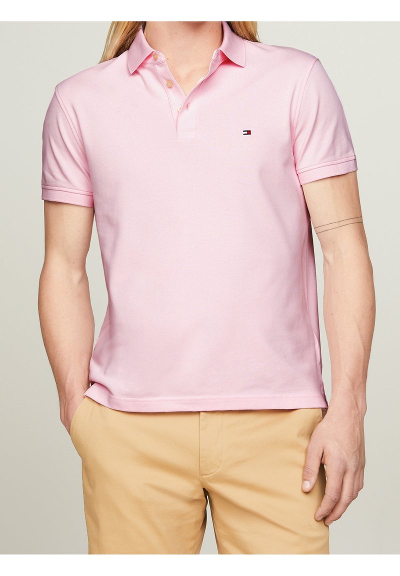 Homme portant un polo rose clair à manches courtes avec un petit logo sur la poitrine et un pantalon beige, mains dans les poches, fond neutre.
