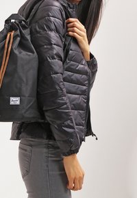 Veste noire gonflée avec fermeture éclair à l'avant et poignets élastiques, associée à un sac à dos noir à cordon de serrage avec un patch logo et des sangles marron.