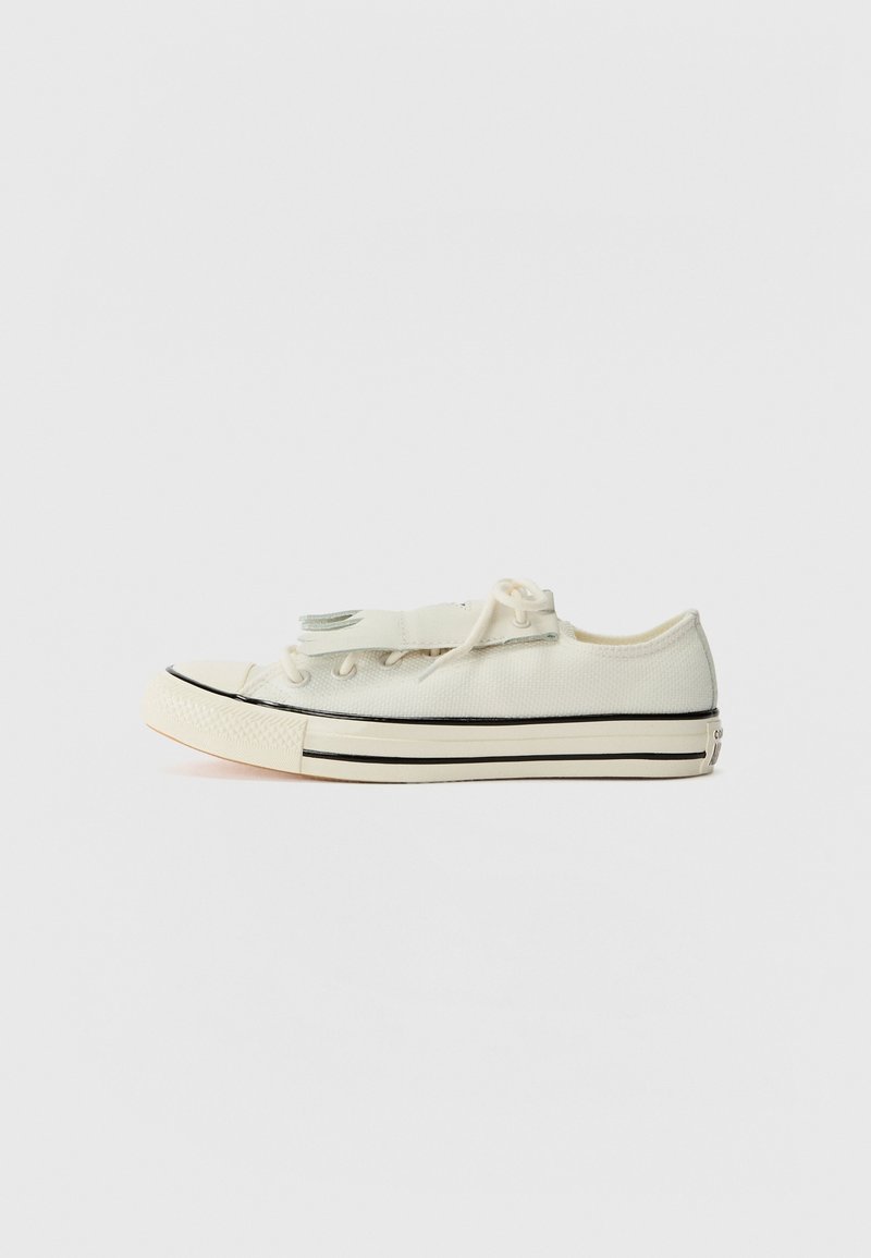 Baskets basses, dessus en toile crème, rayures noires sur la semelle, pointe arrondie avec capuchon en caoutchouc, fermeture à lacets simple, design minimaliste.