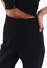 Zwarte op maat gemaakte broeken met een hoge taille, geplooide voorkant en zijzakken. Glad stofoppervlak zichtbaar; de getailleerde snit benadrukt de vorm.