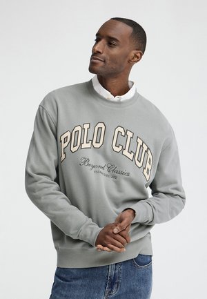Mand iført en lysegrå sweatshirt med teksten "POLO CLUB Beyond Classics", hvid skjorte under og blå jeans, ser til siden.