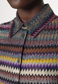 Närbild av en person som bär en skjorta med flerfärgad zigzag-mönstring, svarta knappar och spetskrage.