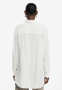 Camicia bianca a maniche lunghe realizzata in tessuto liscio, con una cucitura a yoke sul retro, orlo arrotondato e polsini abbottonati.