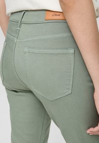 Zielone dżinsy z wysokim stanem, tylnymi kieszeniami z subtelnym przeszyciem oraz brązową skórzaną metką marki blisko pasa. 