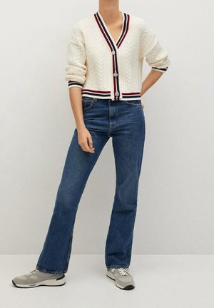 Personne portant un cardigan crème en maille torsadée avec des bordures rouges et bleues, un jean bleu et des baskets grises sur un fond uni.