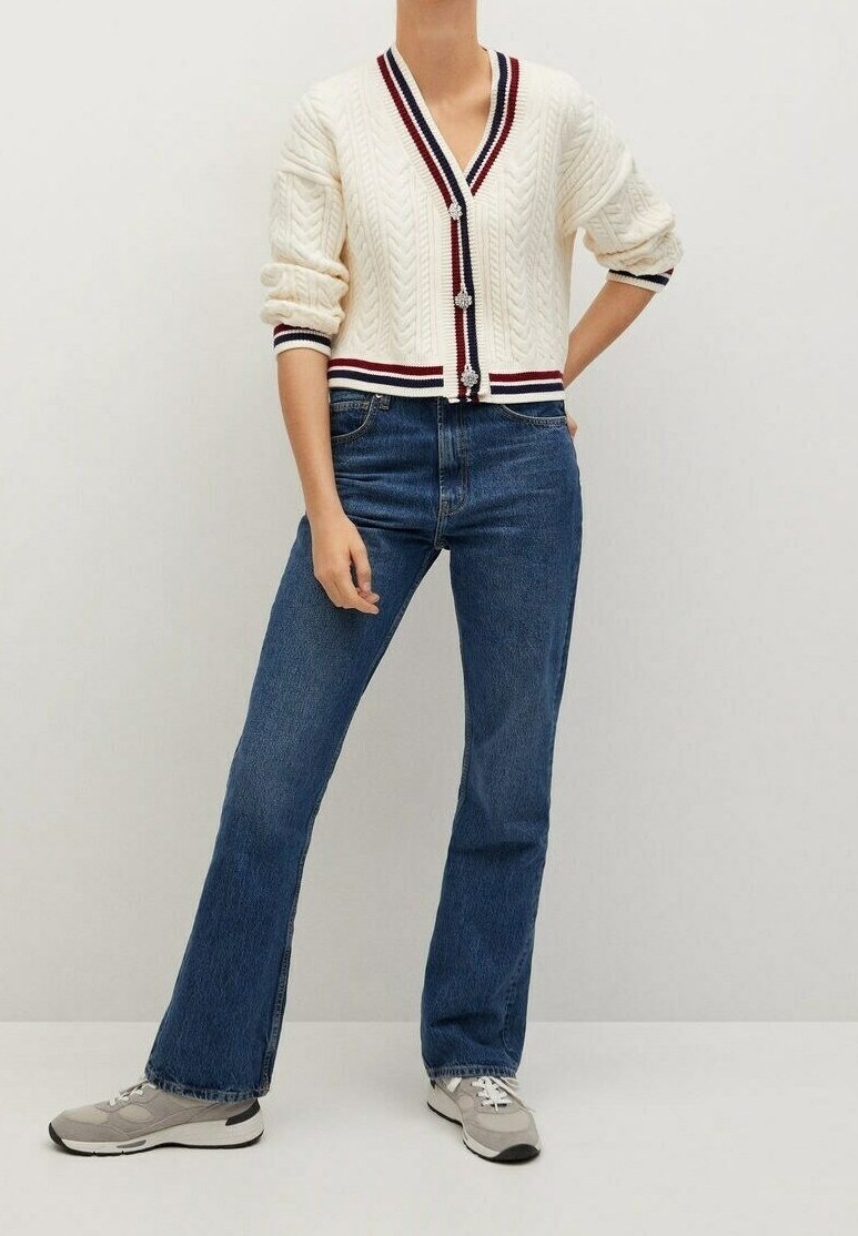 Personne portant un cardigan crème en maille torsadée avec des bordures rouges et bleues, un jean bleu et des baskets grises sur un fond uni.