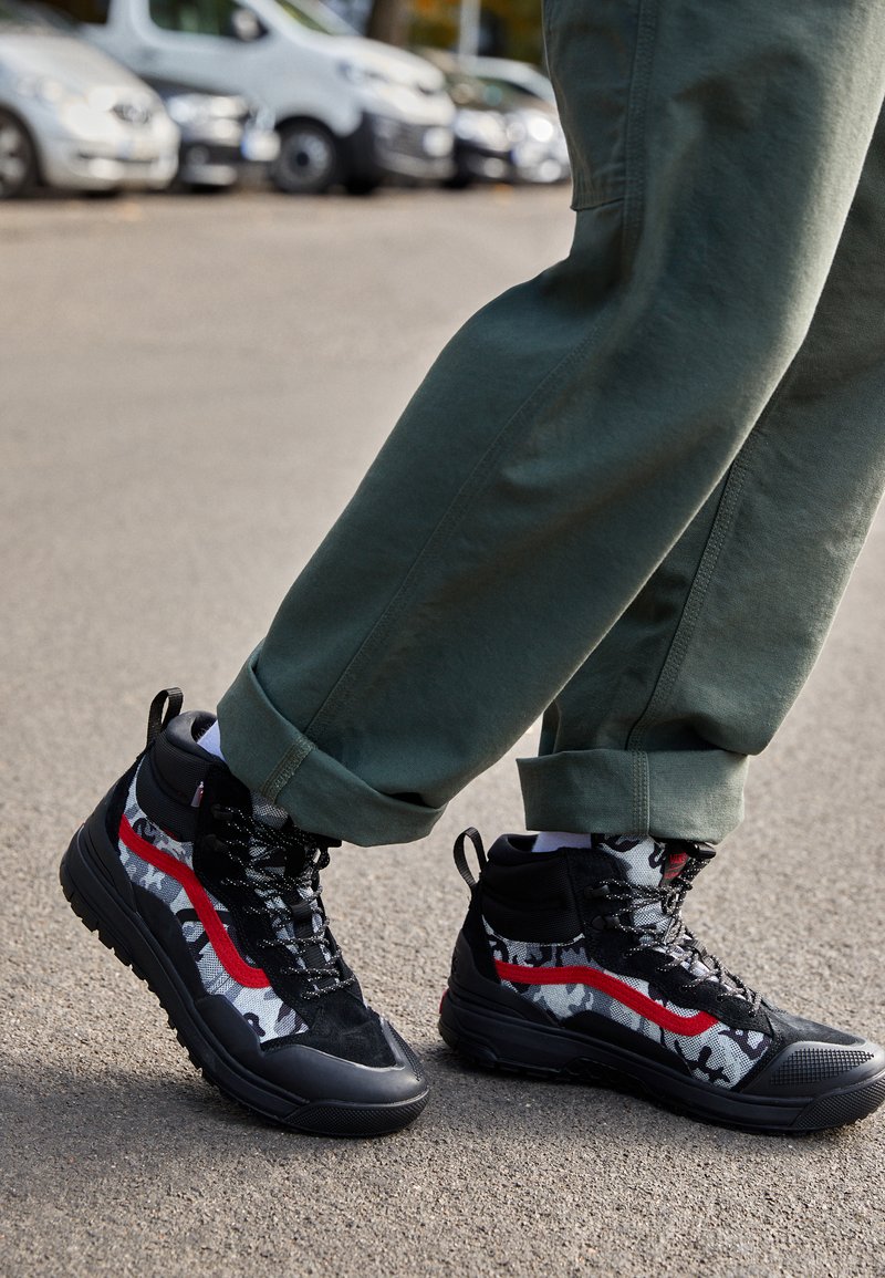 Zapatillas altas en negro y gris con acentos rojos, que presentan un patrón de camuflaje, tela texturizada y suelas de goma para tracción.