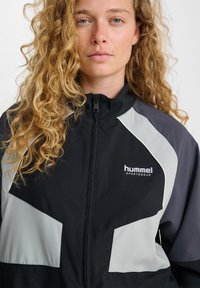 Mladá osoba s kučeravými blond vlasmi, ktorá nosí čiernu a sivú bundu Hummel Sportswear so zipsom vpredu.
