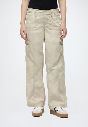 Beige weite Cargohosen mit seitlichen Klappentaschen, getragen mit weißen Sneakern mit schwarzen Details an einer stehenden Person.
