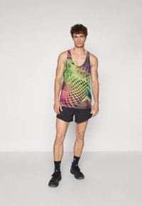 Multifarvet tanktop med abstrakte mønstre i grøn, pink og blå, parret med sorte sports shorts. Model iført sorte sko.