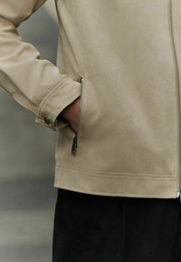 Chaqueta de ante beige con un bolsillo con cremallera. Presenta un cuello alzado y una textura suave. Combinada con una prenda inferior negra.
