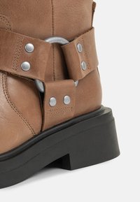 Botte en cuir marron avec des rivets argentés et un anneau métallique au-dessus d'une semelle épaisse en caoutchouc noir.