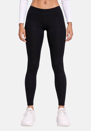 Femme portant un legging noir moulant, un haut blanc à manches longues et des baskets blanches, debout devant un fond uni.