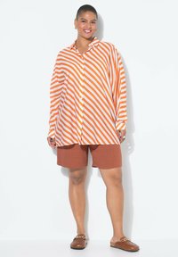 Paski oversize koszula z guzikami w kolorze pomarańczowym i białym, zestawiona z brązowymi szortami i brązowymi butami slip-on z detalem w kształcie łańcucha.