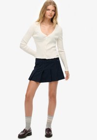 Hvid ribbet cardigan med v-udskæring og knaplukning, parret med en mørkeblå stripet miniskirt. Grå stribede sokker og mørke sko.