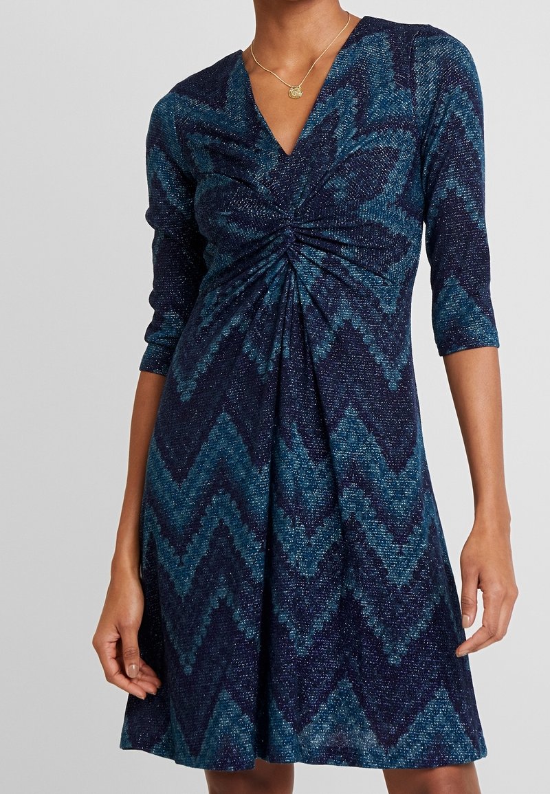 Navy blue and teal zigzag patterned dress with a V-neckline, ruched bodice, three-quarter sleeves, and a flowing A-line skirt.