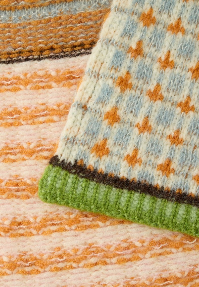 Stickad textil med ett mönster av ljusblå och orange stjärnor, med en grön ribbad kant med struktur, mot en bakgrund med rosa och orange ränder.