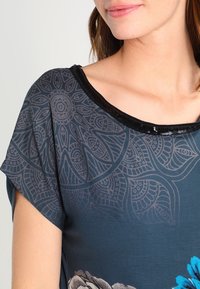 Ciemnoniebieska bluzka z kwiatowym wzorem w odcieniach szarości i niebieskiego. Posiada okrągły dekolt z czarną ozdobną aplikacją z cekinów oraz krótkie rękawy.