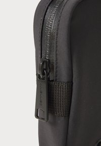Eck eines schwarzen Stoffbeutels mit einem glänzenden Reißverschluss und einem Zugband, strukturierte Oberfläche und einem gewebten Tragegurt. Robustes Design mit nahtlosen Kanten.