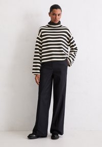 Gestreifter, schwarzer und cremefarbener Strick-Turtleneck-Pullover; oversized Schnitt. Kombiniert mit hochgeschnittenen, weit geschnittenen schwarzen Hosen und klobigen schwarzen Schuhen.