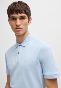 Svijetloplena polo majica od teksturirane tkanine s dvije dugmadi i ugraviranim logom na prsima. Kratki rukavi s klasičnom ovratnikom.