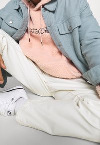 Lichtblauwe button-up jacks, roze fleece hoodie met zwart graphic, crèmekleurige broek en witte sneakers met zwarte accenten. Textuurovergangen zichtbaar.