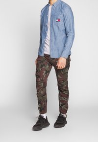 Camicia in denim azzurro chiaro con bottoni sopra una t-shirt grigia, abbinata a pantaloni cargo mimetici verdi e marroni e sneakers nere con calzini bianchi.