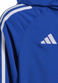 Giacca sportiva blu con strisce bianche sulle spalle e logo ricamato. Tessuto liscio e leggero con un design aderente.
