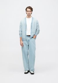 Mlad moški stoji z rokami v svetlo modrem kardiganu, belem T-shirtu, ohlapnih svetlo modrih kavbojkah in črno-belih čevljih na belem ozadju.