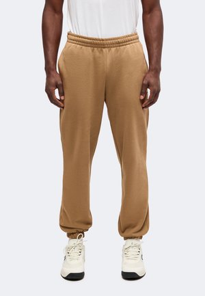 Homme portant un pantalon jogger marron clair avec des poignets élastiques et des baskets blanches, debout devant un fond uni.
