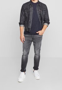 Veste en denim foncé avec les manches retroussées, pull navy, jeans gris slim, et baskets blanches. Tenue décontractée et superposée.