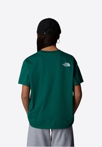 Green cotton t-shirt with a relaxed fit, featuring a small white logo on the upper back. Accompanied by a black cap and gray shorts.