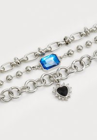 Silver metal chain bracelet featuring three strands: one with a blue gemstone, one with spherical beads, and one with a spiked black heart charm.