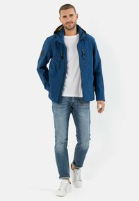 Blaue wasserfeste Jacke mit Kapuze, zwei Reißverschluss-Taschen und einem lebhaften gelben Futter, kombiniert mit hellblauer Jeans und weißen Sneakers.