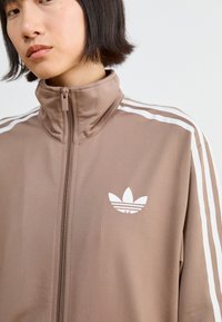 Braune Zip-Jacke mit hohem Kragen, weißen Drei-Streifen-Akzenten auf den Schultern und weißem gesticktem Logo auf der linken Brust. Glatte Textur.