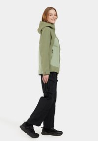Veste à capuche bicolore, verte et gris clair, avec des poches zippées, associée à un pantalon cargo noir et des baskets noires. Manches ajustables incluses.