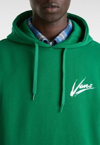 Felpa verde in cotone con una texture morbida, dotata di cordini, un logo frontale bianco e sovrapposta a una camicia a scacchi blu.
