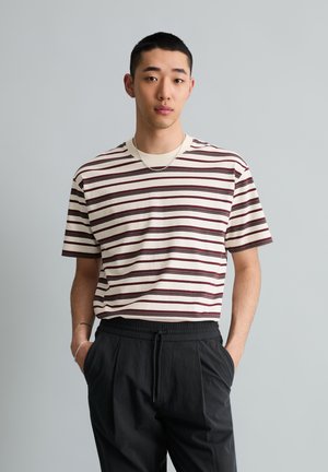 Junger Mann mit kurzen Haaren, der ein weiß-braun gestreiftes T-Shirt und schwarze Hosen trägt, steht mit den Händen in den Taschen vor einem einfarbigen Hintergrund.