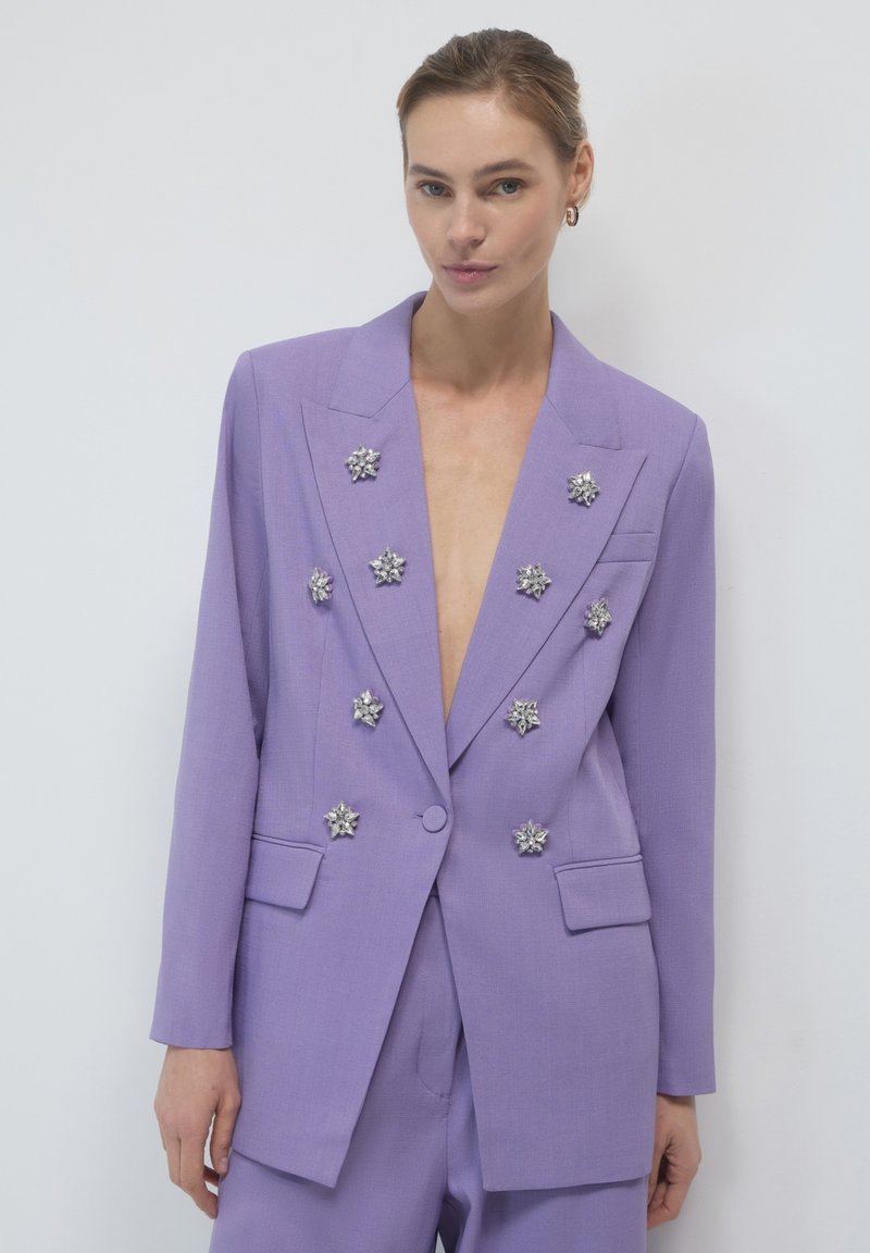 Blazer lavanda con scollatura a V profonda e decorazioni floreali argentate. Forma sartoriale, due tasche frontali e tessuto leggero e testurizzato.