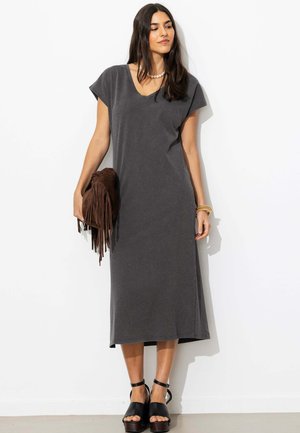 Femme portant une robe midi gris charbon et des sandales compensées noires, tenant un sac à main marron à franges, debout contre un mur blanc.