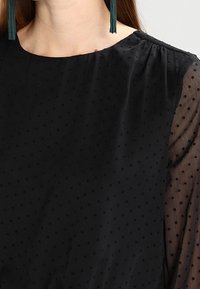 Vrouw in een zwarte doorzichtige blouse met kleine stippen en lange groene franjeringen, waarbij nek- en schouderdetails worden getoond.