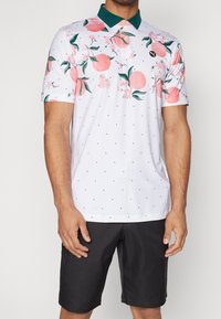 Krátký rukáv polo tričko v bílé barvě s růžovými ovocnými a květinovými vzory, zeleným límcem a černými puntíky, spárované s černými šortkami.