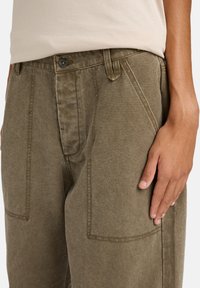 Grüne Baumwollhose mit strukturiertem Finish, mit Knopfverschluss, Seitentaschen und sichtbaren Nähten entlang der Säume.