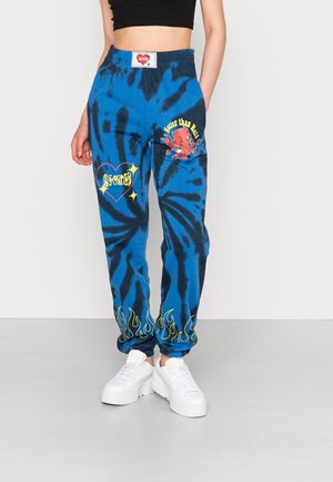 Blauw-zwarte tie-dye sweatpants met gele vlamprint op de boorden, rode duivelgrafiek en gele tekstpatches, gedragen met witte sneakers.