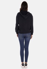 Felpa nera con cappuccio regolabile, maniche a costine e una vestibilità rilassata, abbinata a jeans blu aderenti e sneakers bianche.