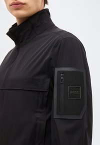 Κοντινό πλάνο ενός ατόμου που φοράει μαύρο μπουφάν με ψηλό γιακά και ψηφιακή ετικέτα με το λογότυπο BOSS με φερμουάρ στο επάνω αριστερό μανίκι.