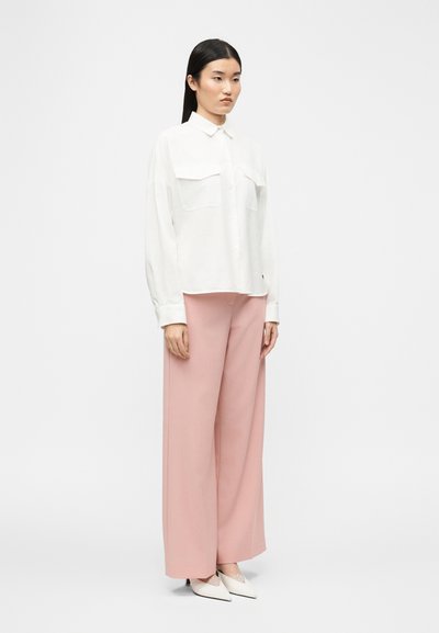 Γυναίκα που στέκεται φορώντας ένα λευκό μακρυμάνικο πουκάμισο με κουμπιά, ροζ παντελόνι με φαρδύ πόδι, και λευκές μυτερές γόβες, κοιτάζοντας ελαφρώς προς τα δεξιά.